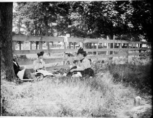 Black and white photo of 2 women and 1 man eating a picnic meal next to a fence and under a tree. Dated 1899