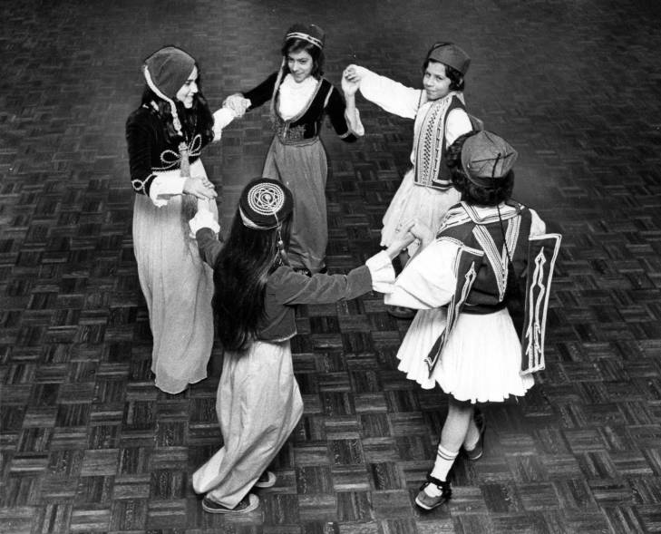a group of 5 women dressed in traditional Greek folkwear hold hands in a circle while dancing
