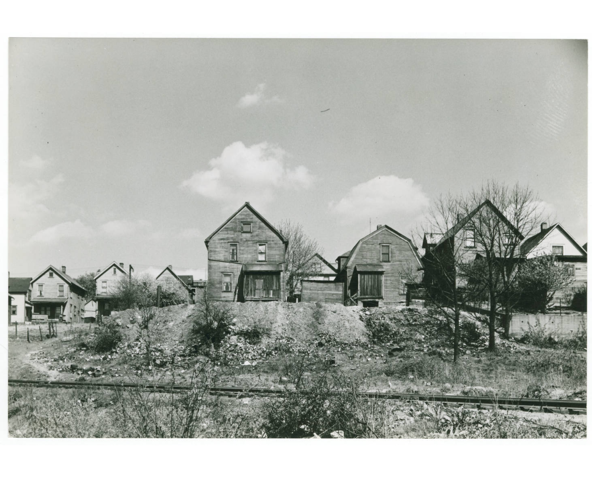 black and white photo of a row of houses with greenery in front of them and a few clouds in the sky above