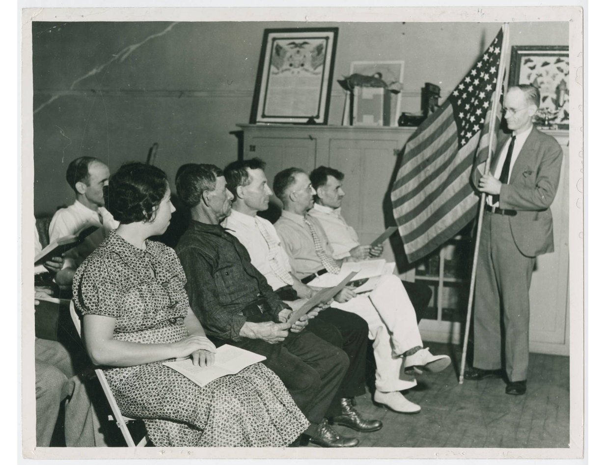 Black and white photo of 6-7 people seated and listening to a man before them speak