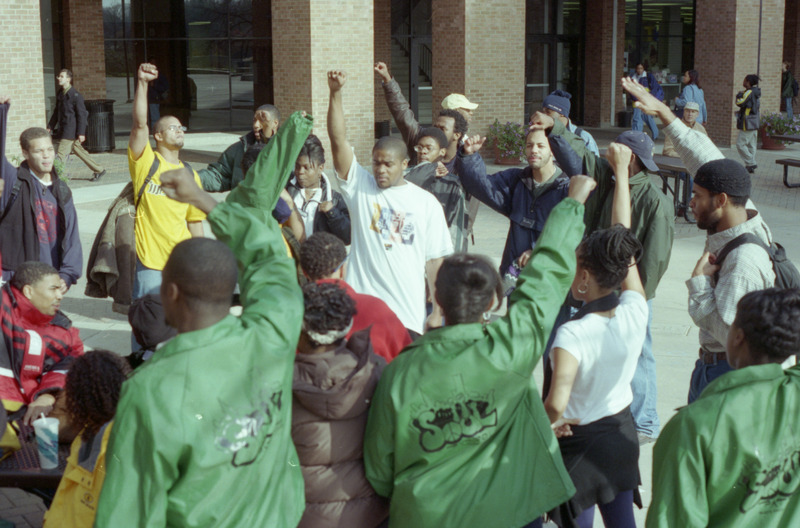 Color photograph of a group of African American men standing in a group outside with their fists raised in the air